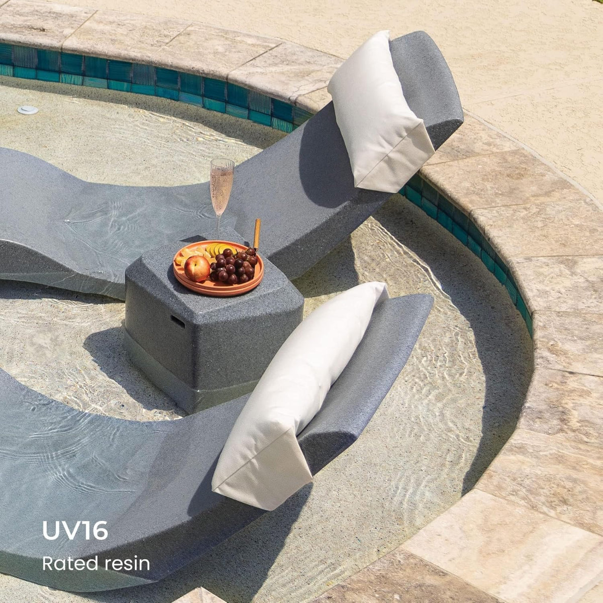 Two gray loungers with white cushions are partially submerged in a circular pool. A small table between them holds a glass of champagne and a bowl of fruit. Text on the image reads UV16 Rated resin. The pool is surrounded by a stone edge.