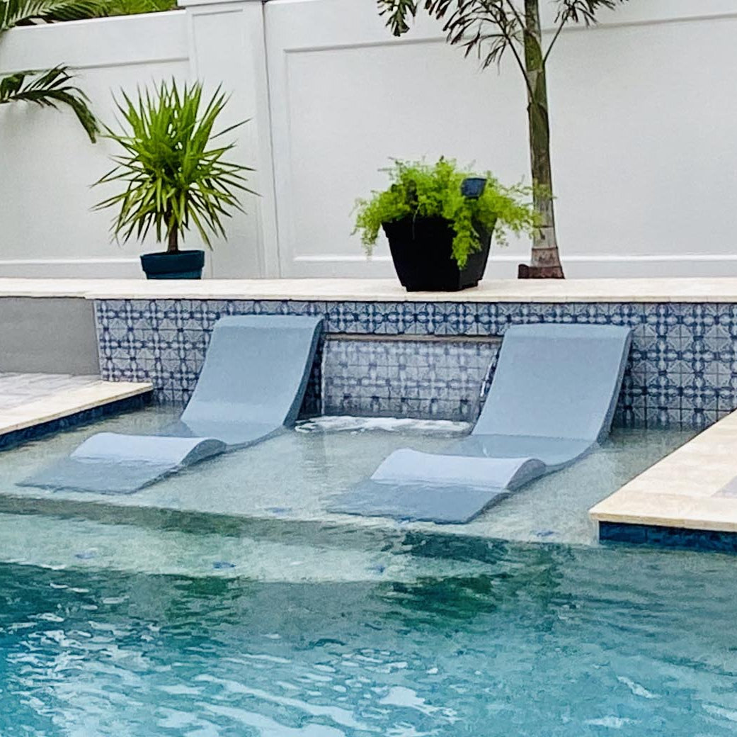 Two Aqua Outdoors Chaise Lounge Set chairs in light blue are partially submerged on a pool riser, with potted plants and a white fence behind them. The modern setup is ideal for relaxing at the perfect water depth.
