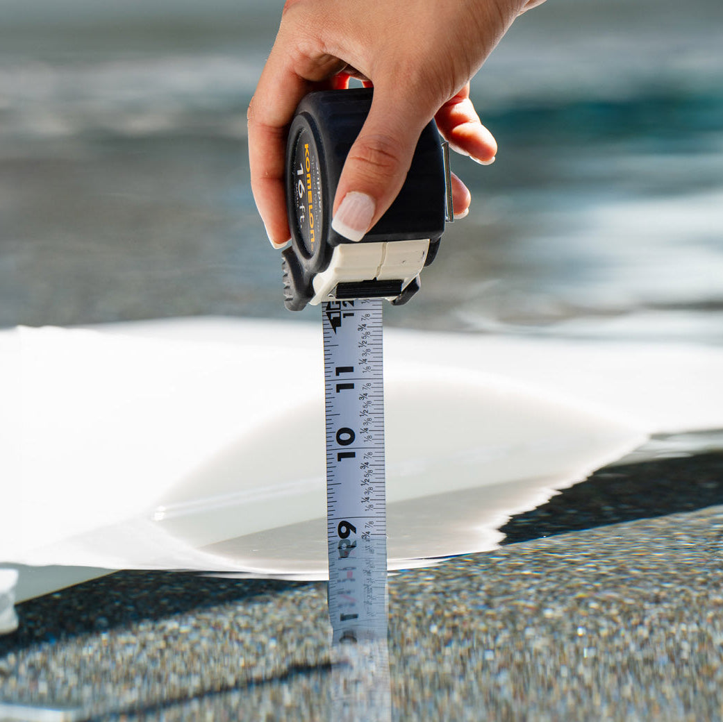 A hand uses a tape measure to check water depth under the Chaise Lounge Set by Aqua Outdoors, with the tool upright on a white surface.