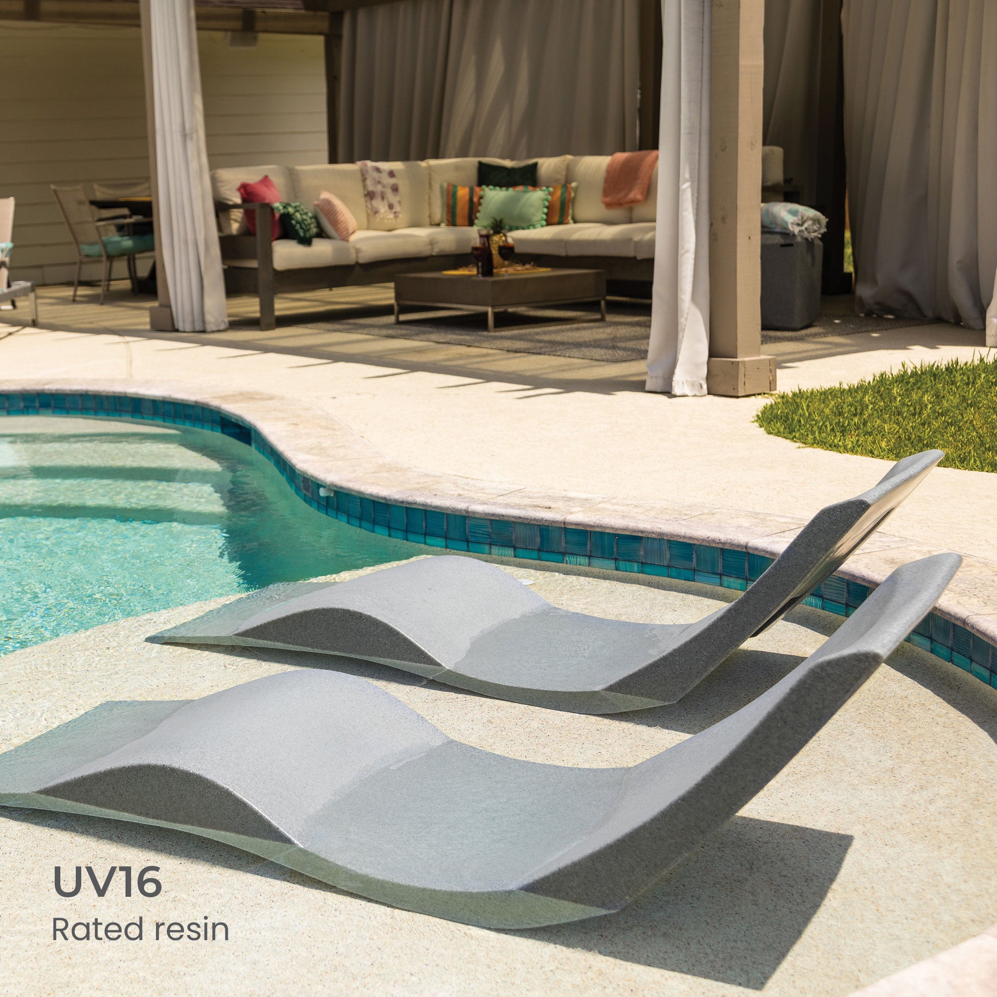 Two modern lounge chairs partially submerged in a pool with a tiled edge. In the background, theres a covered patio with a sectional sofa, cushions, and a small table. Grass borders the pool area. Text reads, UV16 Rated resin.