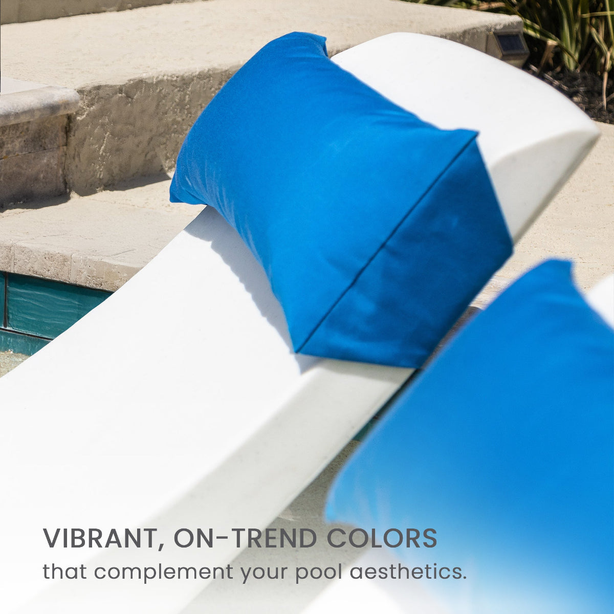 Two blue outdoor pillows are placed on white reclining pool chairs beside a turquoise-tiled pool. A stone surface and sand are visible in the background. Text reads, Vibrant, on-trend colors that complement your pool aesthetics.