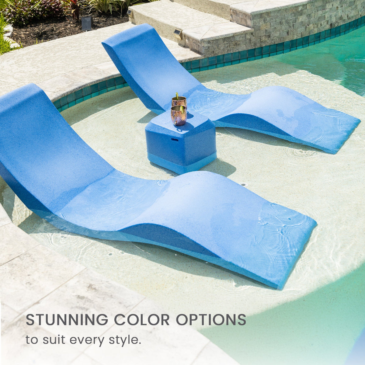 Two blue, wavy chaise lounges are partially submerged in a shallow pool area. A small blue table between them holds a drink. Text reads Stunning color options to suit every style. The setting is sunny, with a stone pool edge and some greenery.