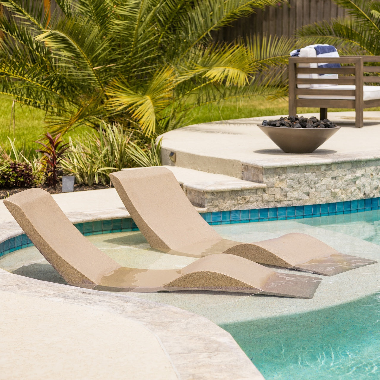 Two beige lounge chairs partially submerged in a pool next to a stone edge. A round fire pit is near the chairs, and a wooden chair with a striped towel is in the background. Palm trees and greenery surround the pool area.