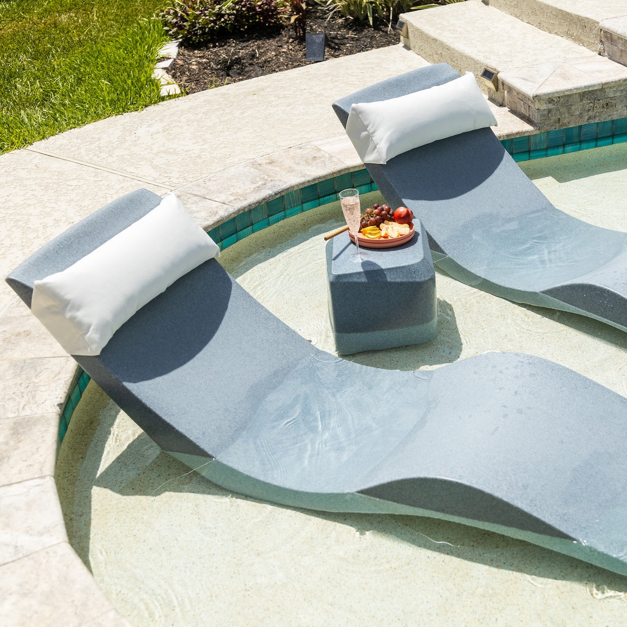 Two modern lounge chairs with white cushions are partially submerged in a shallow pool. A small table between them holds a plate of dessert and a glass of champagne. The pool area is surrounded by grass and a stone patio.